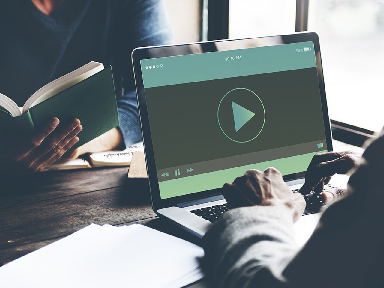 Bilgisayar ekranında görünen video oynatma görüntüsü ve bilgisayarın arka planında görünen kitap okuyan adam