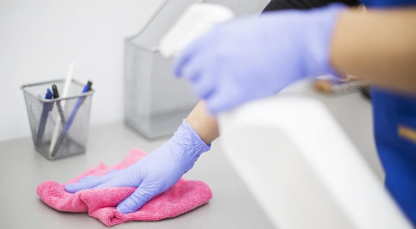 Pembe bir bez ve dezenfektan ile iş yerini temizleyen eldivenli bir kişi